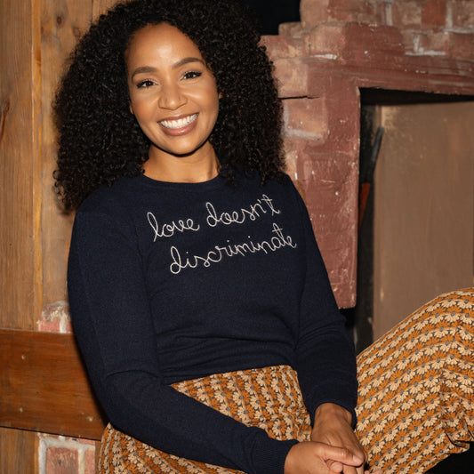 Woman wearing a navy sweater with text, sitting on a wooden bench against a brick wall.